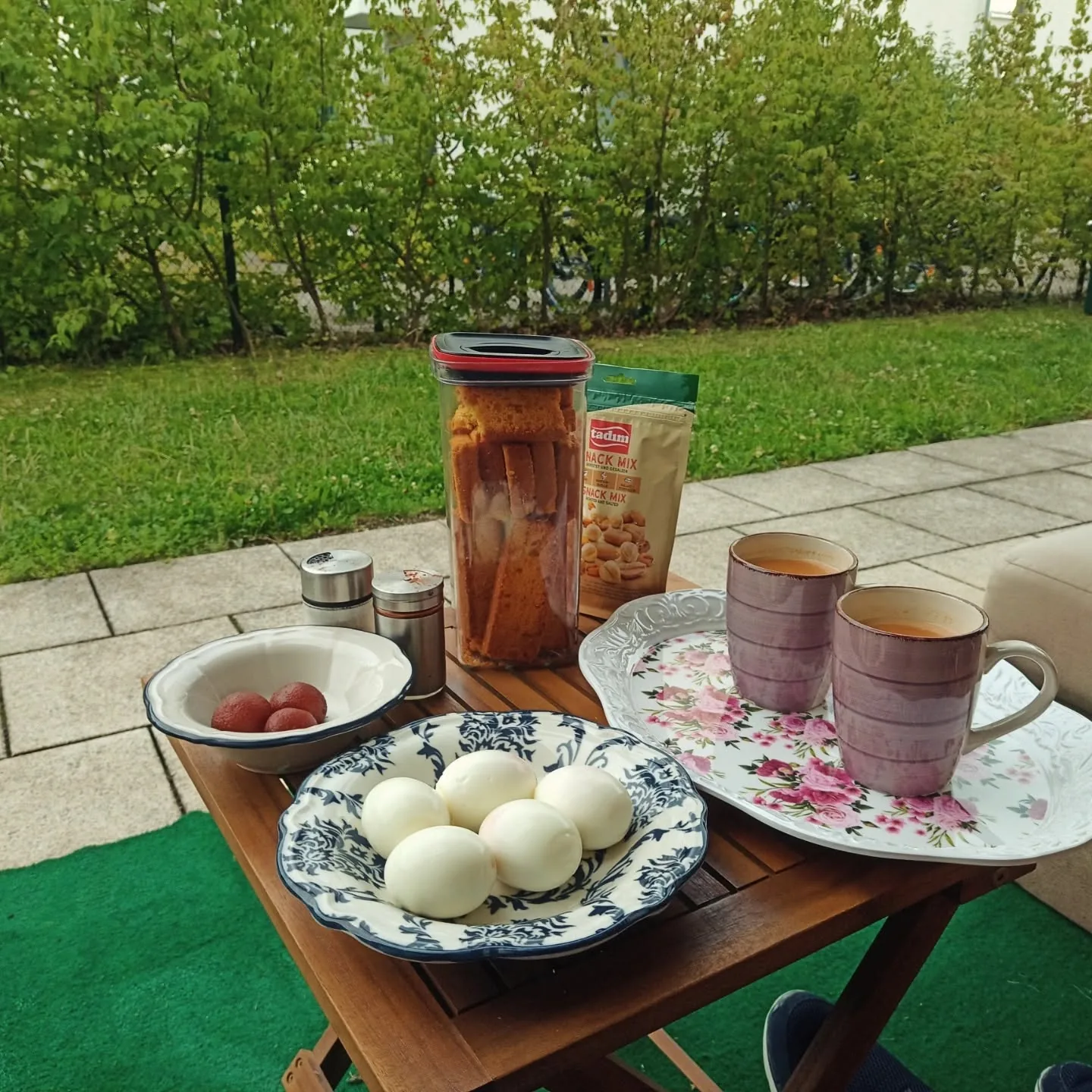 An evening tea spread on a folding table in a German garden: two cups of chai, a bowl of eggs, sweets, and a packet of snacks, with a green hedge behind.