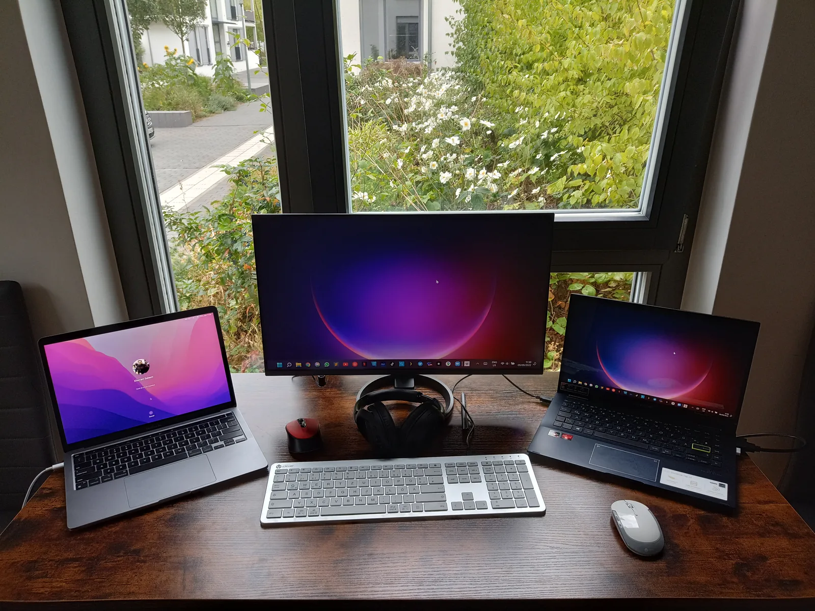 The desk – MacBook, main monitor, and an Asus laptop side by side at the window